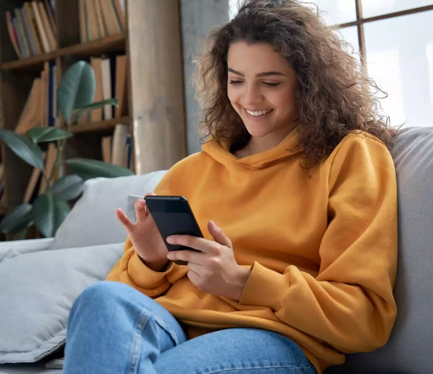 Eine junge Frau sitz auf dem Sofa und schaut lächelnd auf das Smartphone in ihrer Hand, mit dem sie interagiert