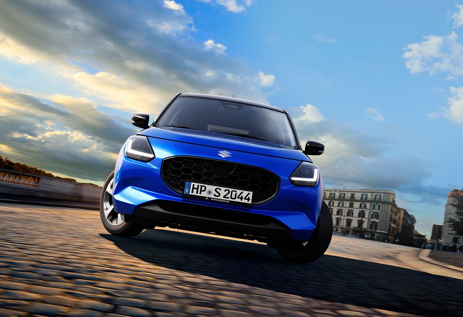 Blauer Suzuki Swift 1.2 DUALJET HYBRID Comfort+ von schräg unten vor City-Setting auf Kopfsteinpflaster