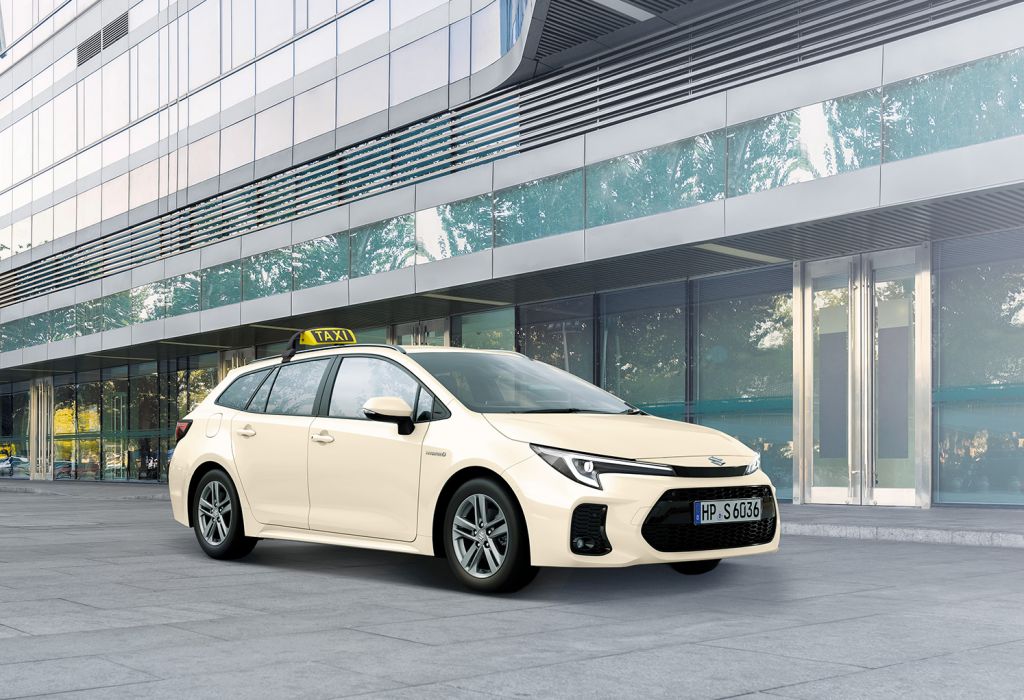 Beiger Suzuki Swace 1.8 HYBRID CVT Comfort+ mit Taxischild vor Glasfront eines Gebäudes in Front-Seitenansicht