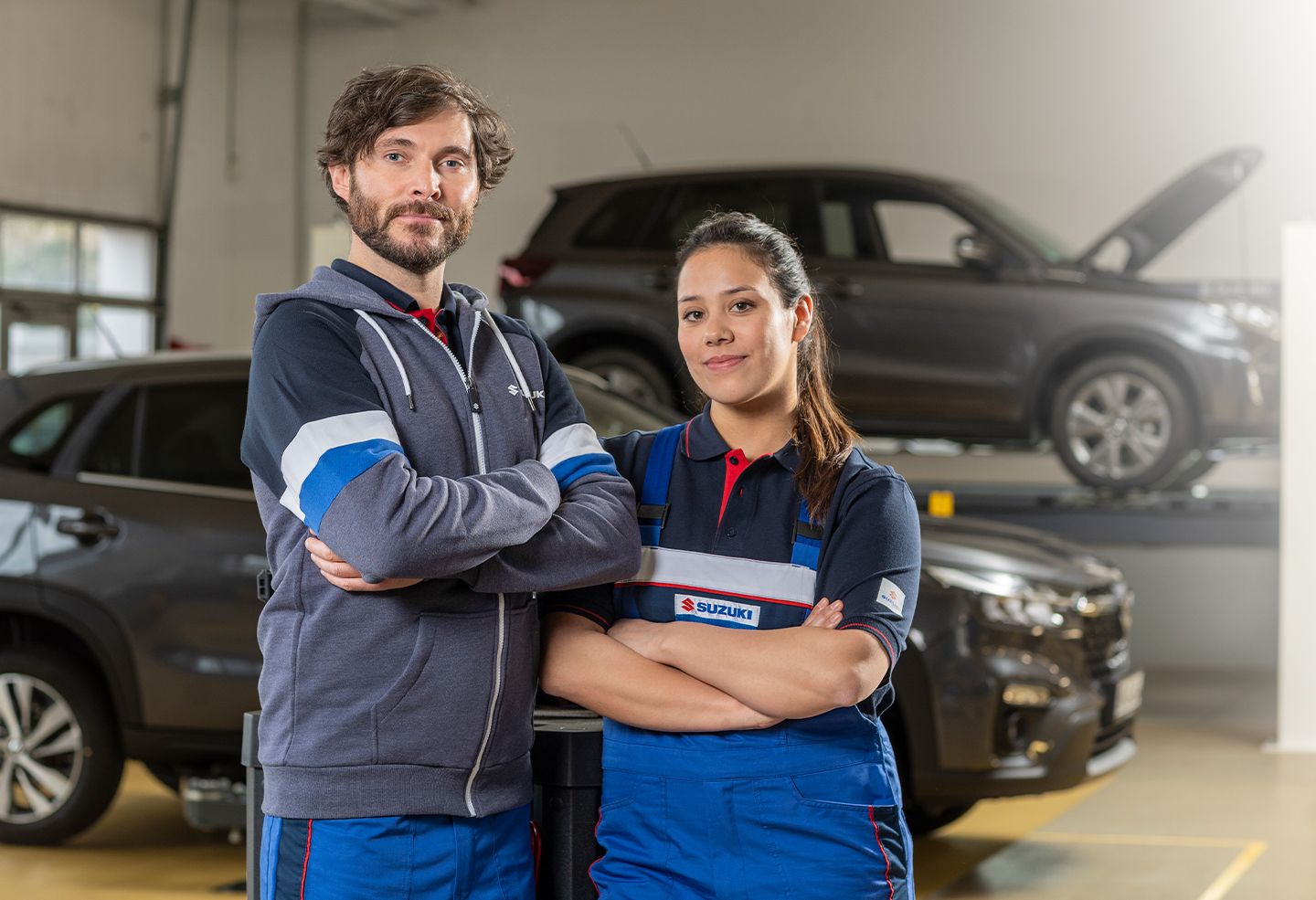 Zwei freundlich lächelnde Kfz-Mechatroniker:innen stehen in der Werkstatt vor zwei Suzuki Modellen