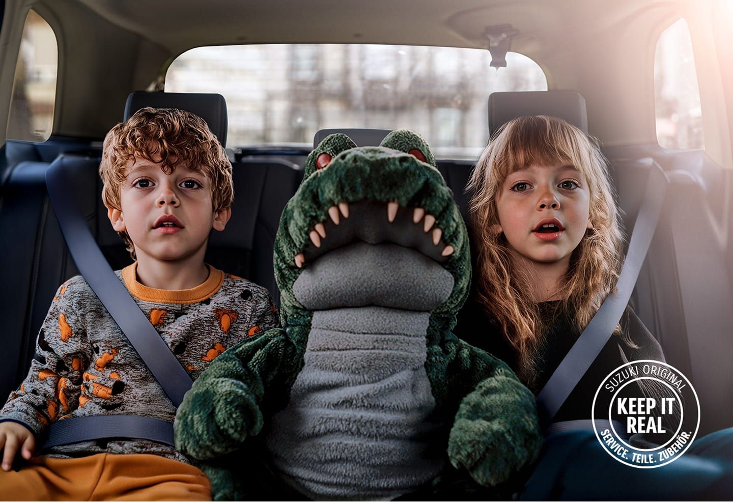Mädchen und Junge angeschnallt auf der Rückbank eines Fahrzeugs, zwischen ihnen ein Stoffkrokodil, unten rechts das Keep it real-Logo