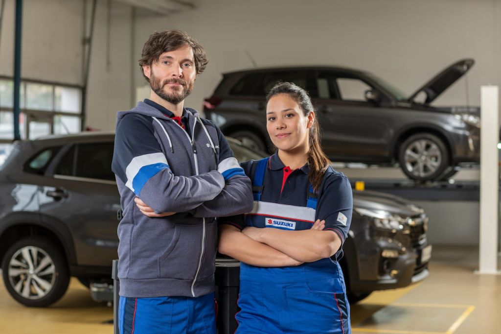 Zwei freundlich lächelnde Kfz-Mechatroniker:innen stehen in der Werkstatt vor zwei Suzuki Modellen