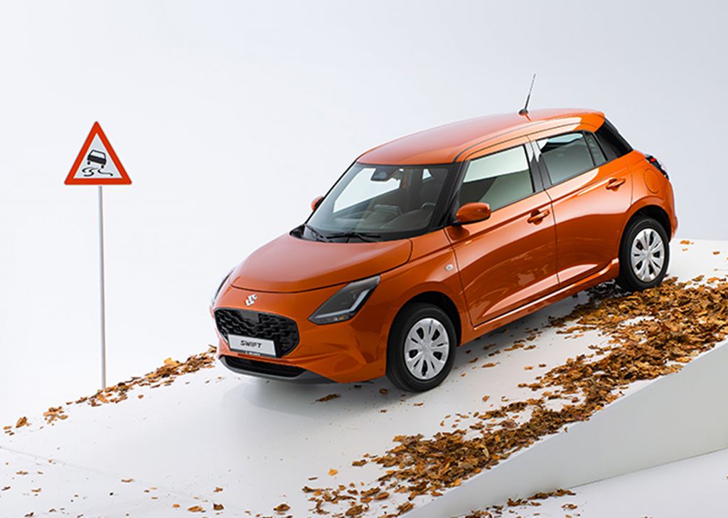 Der Suzuki Swift in orange steht auf einer weißen Rampe mitsamt Laub, daneben ein Verkehrswarnschild für Rutschgefahr.