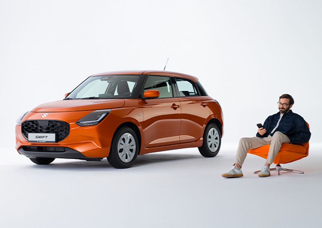 Ein Mann in legerer Kleidung mit Bart und Brille sitzt in einem orangefarbenen Sessel neben einem Suzuki Swift in Orange, er blickt lächelnd auf das Display eines Smartphones in seiner Hand.