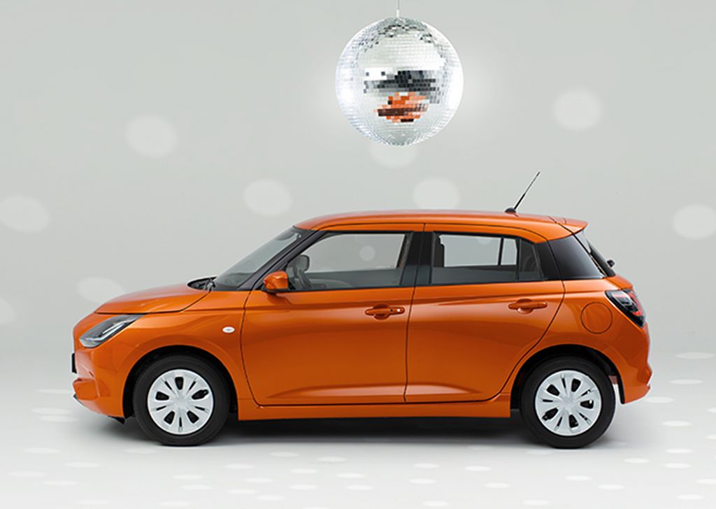 Der Suzuki Swift in orange steht vor einem neutralen weißen Hintergrund, über ihm glitzert eine Discokugel und wirft gespiegelte Lichter an die Wand und auf den Boden.